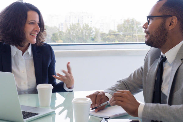 A professionally dressed white woman speaking with a professionally dressed dark-skinned man in front of a laptop.