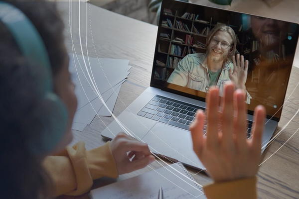 Two people using video conferencing to collaborate. One in the foreground wearing headphones and waving. One on the laptop screen waving back.